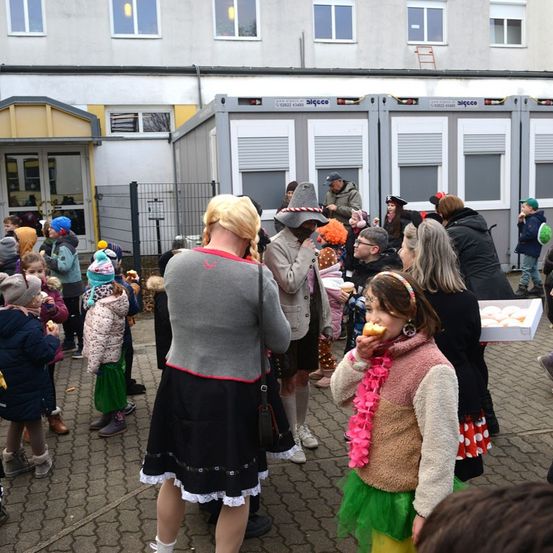 Eine Menschenmenge, einschließlich Kinder, versammelt sich im Freien. Einige essen Donuts, andere tragen Kostüme. Eine Frau in einem grauen Top und schwarzer Rock hält eine Handtasche.