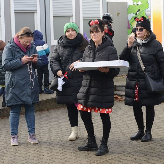 Vier Frauen in Winterkleidung stehen draußen, eine hält eine Pizzaschachtel. Sie stehen auf einem Ziegelpfad mit einem Gebäude im Hintergrund. Eine Frau trägt Minnie-Mouse-Ohren.