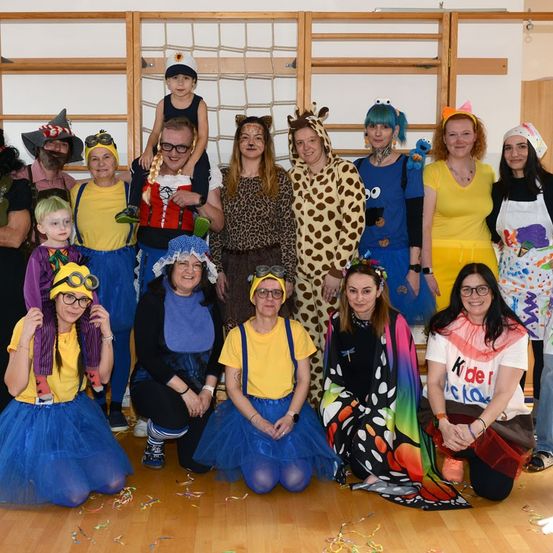 Eine Gruppe von Menschen in Kostümen posiert für ein Foto. Dazu gehören ein Kind auf den Schultern eines Mannes, eine Frau in einem Giraffenkostüm und andere in verschiedenen bunten Kostümen.