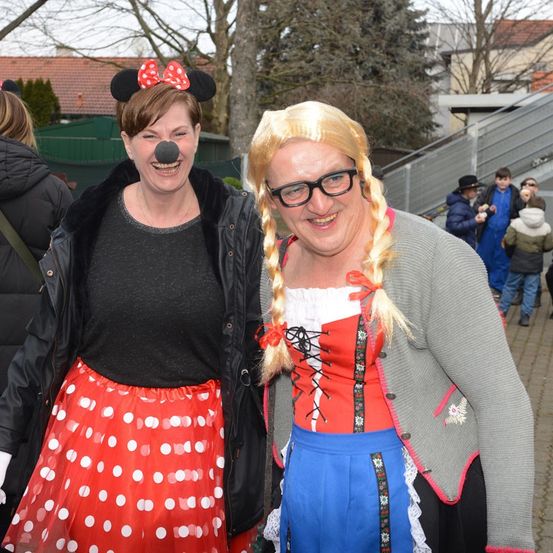 Zwei Frauen in Kostümen lächeln für die Kamera, eine in einer Minnie-Mouse-Verkleidung und die andere in einem traditionellen bayerischen Kleid. Sie stehen im Freien, mit einem Baum und Treppen im Hintergrund.