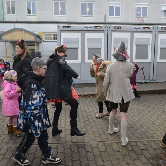 Eine Gruppe von Menschen, einschließlich Kinder, versammelt sich auf einer gepflasterten Oberfläche vor einem Gebäude. Einige von ihnen tragen Kostüme.