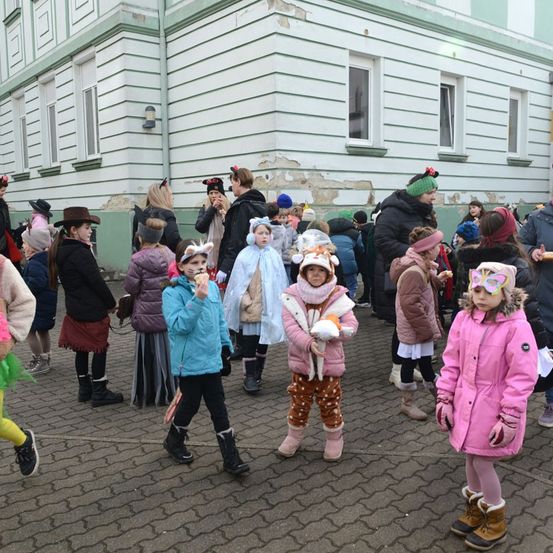 Eine Gruppe von Kindern in verschiedenen Kostümen steht vor einem Gebäude. Einige tragen Hüte, andere Stiefel. Ein Mädchen hält eine Orange.