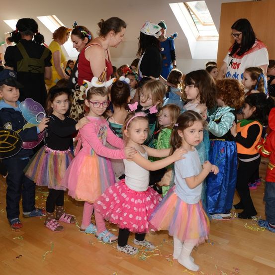 Eine Gruppe von Kindern in Kostümen und Tüllröcken ist drinnen versammelt, Erwachsene schauen zu. Einige Kinder halten Requisiten.