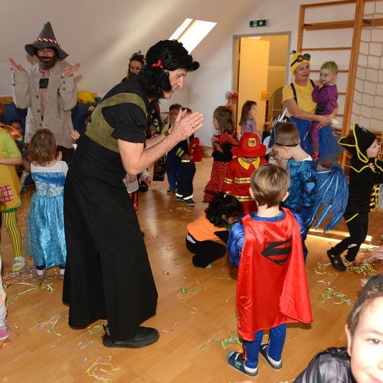 Eine Gruppe von Kindern in Kostümen tanzt in einem Raum, einige mit einem Clown und einem Erwachsenen in einem Piratenkostüm. Ein Feuerwehrmann und ein Superheld sind ebenfalls anwesend.