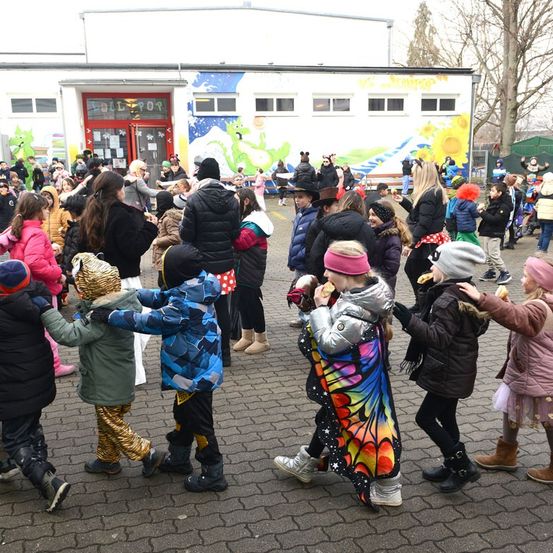 Eine Gruppe von Kindern tanzt vor einem Gebäude mit bunten Wandgemälden. Einige sind in Kostümen gekleidet, andere halten Essen in der Hand.