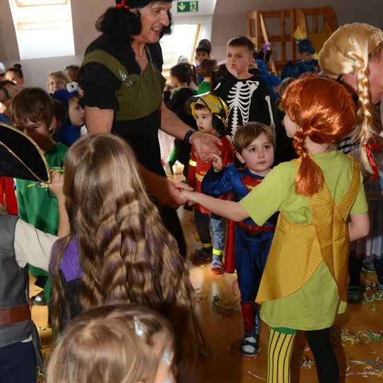Eine Gruppe von Kindern in Kostümen ist um eine Frau herum versammelt. Sie sind drinnen, möglicherweise auf einer Kostümparty. Einige Kinder halten die Hand der Frau.