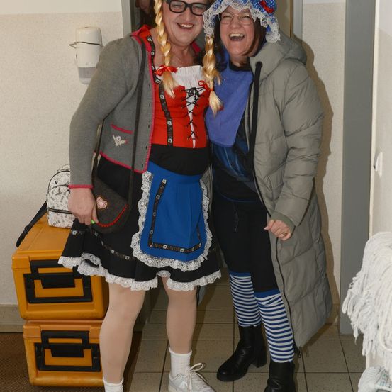 Zwei Frauen in einem Raum, eine in traditioneller Kleidung mit Haube, die andere in einer gefütterten Jacke und gestreiften Socken, beide lächeln für die Kamera.