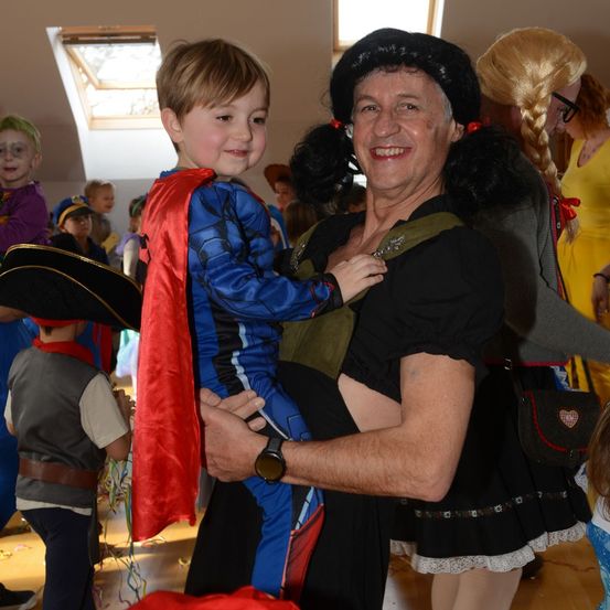 Ein erwachsener Mann in einem schwarzen Kleid und einer Haube trägt einen lächelnden Jungen in einem Superheldenkostüm. Mehrere Kinder in verschiedenen Kostümen umgeben sie, und eine Person mit blondem Haar steht hinter ihnen.