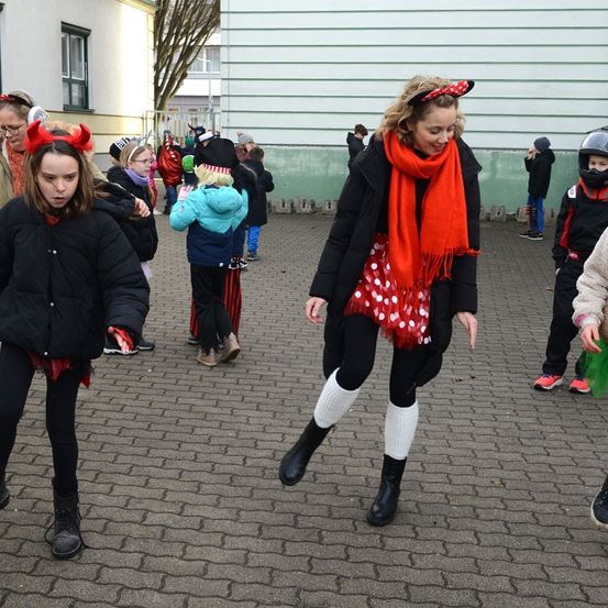 Gruppe von Kindern und Erwachsenen in Winterkleidung, einige in Kostümen, gehen auf einem Ziegelweg. Eine Frau mit rotem Schal und Minnie-Mäuse-Ohren führt.
