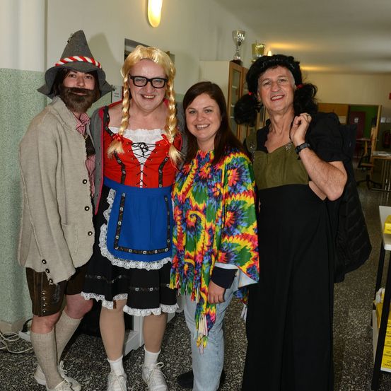 Vier Menschen in Kostümen stehen zusammen in einem Flur. Ein Mann in traditioneller deutscher Kleidung, eine Frau in einem bayerischen Kleid und zwei weitere in bunten und dunklen Kleidungsstücken. Sie lächeln.