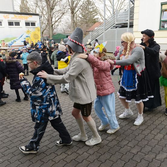 Eine Gruppe von Kindern und Erwachsenen tanzt im Kreis draußen. Einige tragen Hüte und Kostüme. Ein Kind hält ein Stück Essen in der Hand.