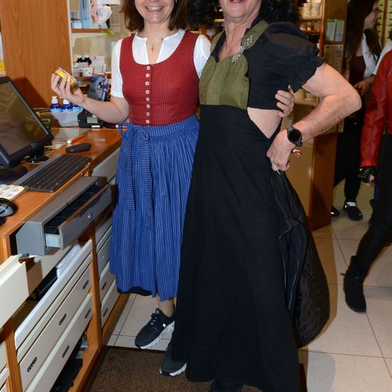 Zwei Frauen in traditionellen bayerischen Kostümen posieren für ein Foto in einem Geschäft. Eine hält eine Dose. Ein Computer, Tastatur und Schubladen sind hinter ihnen.