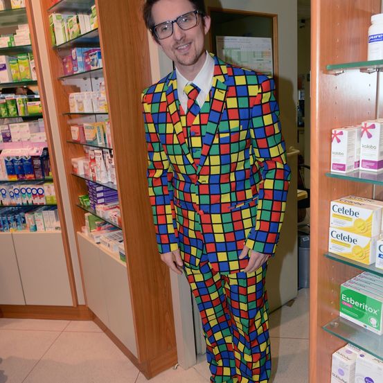 Ein Mann in einem bunten Anzug steht in einer Apotheke, umgeben von Regalen mit Medizin.