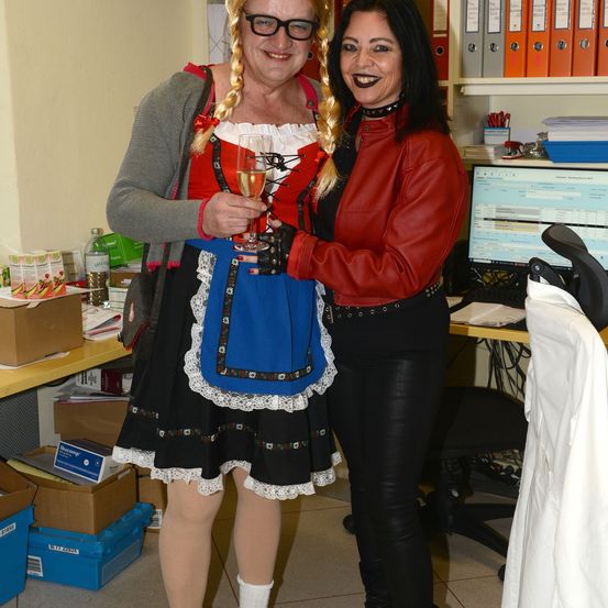 Zwei Frauen stehen nah beieinander in einem Büro, eine trägt ein traditionelles deutsches Kostüm und hält ein Weinglas, die andere eine rote Jacke.