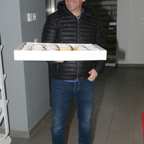 Ein Mann in einer schwarzen Jacke und blauen Jeans hält eine Schachtel mit Donuts in einem Flur.
