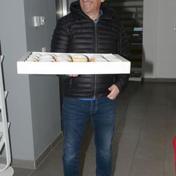 A man in a black jacket and blue jeans is holding a box of donuts, standing in a hallway.