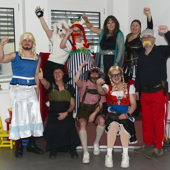 Eine Gruppe von Menschen in verschiedenen Kostümen posiert für ein Foto. Einige tragen Helme und Brillen, andere haben Perücken und Stiefel. Eine Person gibt einen Daumen hoch.