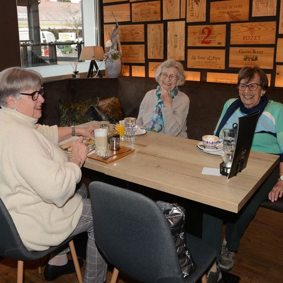 Drei Frauen sitzen in einem Café an einem Tisch und lächeln. Die Frau auf der linken Seite hält ein Glas Saft, während die anderen Kaffeetassen haben. Dahinter befindet sich eine Holzwand mit verschiedenen Etiketten.