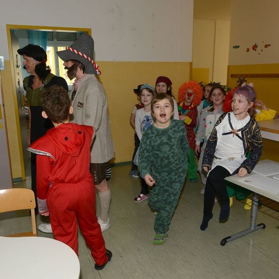 Kinder in Kostümen stehen und sitzen in einem Raum, ein Clown ist dabei. Ein Mann und eine Frau mit Hüten stehen vor der Tür.