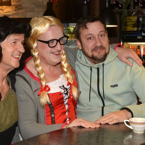 Drei Personen sitzen an einem Tisch; eine Frau mit blonden Haaren und einer Brille, ein Mann mit Bart und eine weitere Frau in einem Kostüm. Sie lächeln, eine Frau hält eine Tasse.