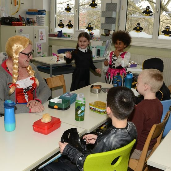 Ein Klassenzimmer mit Kindern, einer Frau und Brotdosen auf dem Tisch. Im Hintergrund tanzen zwei Mädchen.