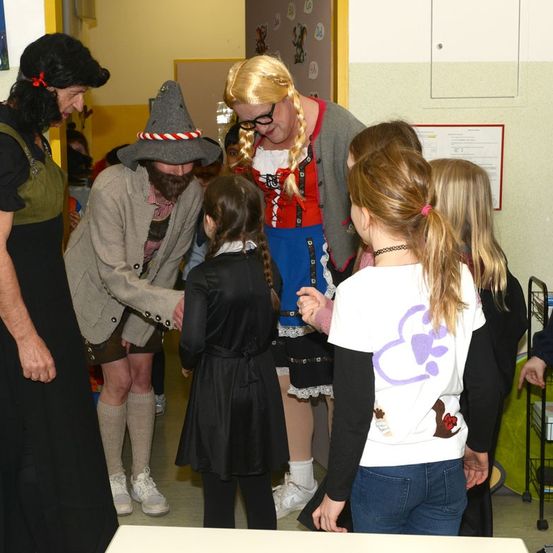 Mehrere Menschen in Kostümen stehen in einem Raum, vermutlich in einer Schule. Eine Frau mit blonden Haaren trägt ein traditionelles deutsches Kostüm, und ein Mann in einem Hut spricht mit einem Mädchen.