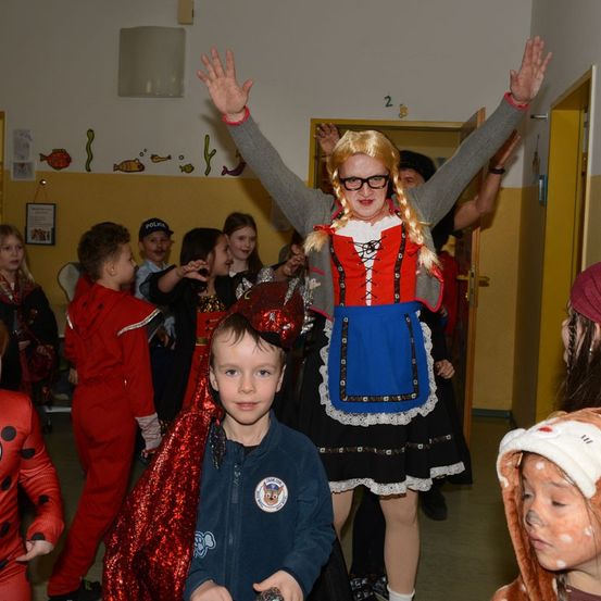 Eine Frau in traditioneller Kleidung hebt die Arme, während ein Kind zuschaut, mit anderen Kindern in Kostümen in der Nähe.