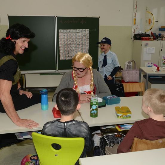 Eine Frau in einem Kostüm sitzt mit drei Kindern an einem Tisch. Ein Lehrer in Uniform steht hinter dem Tisch. Im Hintergrund befindet sich eine Tafel mit Zahlen und ein Diagramm.