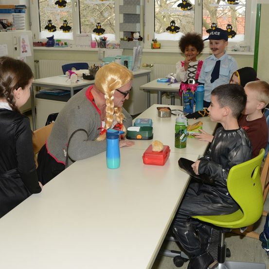 Eine Frau mit blonden Haaren sitzt mit Kindern in Kostümen an einem Tisch. Sie sitzen an einem langen Tisch in einem Klassenzimmer mit Fenstern dahinter.