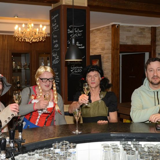 Vier Personen in traditionellen Kostümen stoßen mit Weinläsern an einer Bar an. Sie sitzen um einen Tresen herum, vor ihnen ein Menübrett.