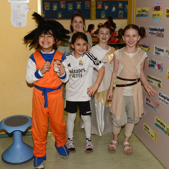 Fünf Kinder in Kostümen posieren für ein Foto in einem Klassenzimmer, eins hält eine Karte. Der Junge trägt ein Dragon Ball Z-Outfit, das Mädchen neben ihm ein Star Wars-Kostüm.