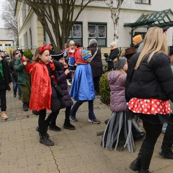 Eine Gruppe von Kindern und Erwachsenen in verschiedenen Kostümen läuft auf einem gepflasterten Gehweg vor einem Gebäude.