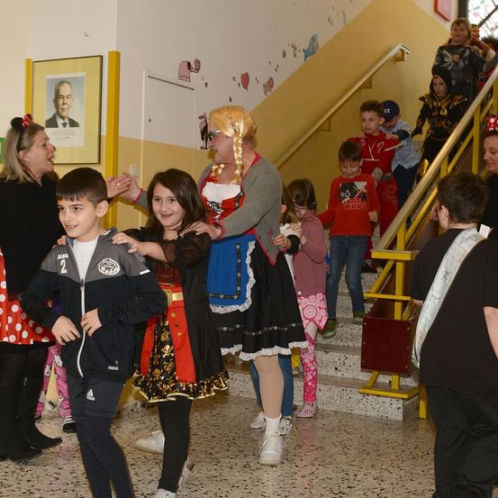 Eine Gruppe von Kindern und Erwachsenen versammelt sich in einem Flur. Einige sind in Kostümen gekleidet, während andere normale Kleidung tragen. Eine Frau in einem Kostüm hilft einem jungen Mädchen und Jungen auf den Treppen.