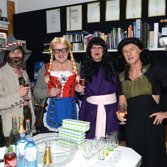 Vier Erwachsene in Kostümen posieren für ein Foto in einem Raum mit Flaschen und Gläsern auf einem Tisch. Ein Mann mit Hut hält ein Glas Wein.
