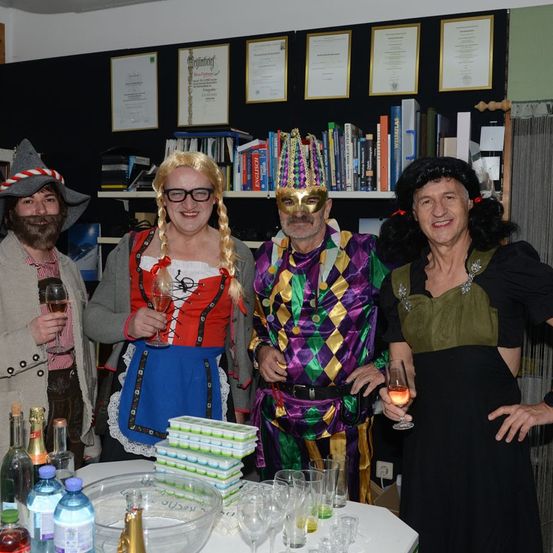Vier Erwachsene in Kostümen stehen bei einer Party zusammen. Zwei Männer tragen Hüte, einer hat einen Bart. Eine Frau in einem roten Kleid lächelt und ein Mann in bunter Kleidung hält ein Glas. Ein Tisch hat Flaschen, Weingläser und eine Schüssel.