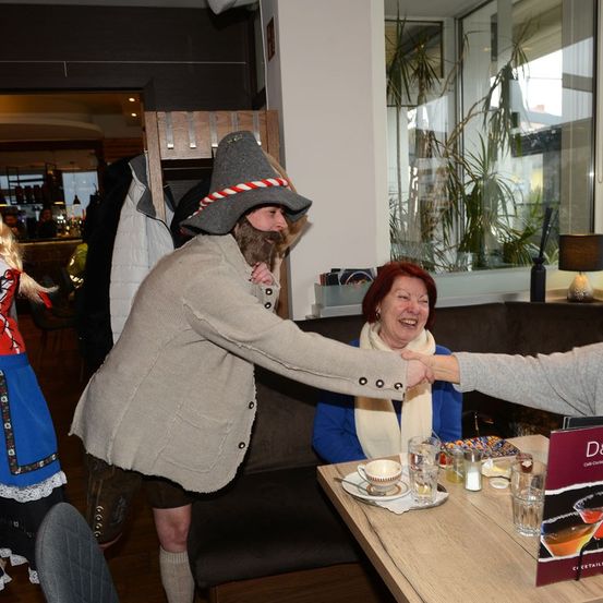 Ein Mann in traditioneller Kleidung schüttelt die Hand einer Frau in einem Café. Vor ihnen steht ein Tisch mit Speisekarte und Getränken. Eine Frau in traditioneller Kleidung steht in der Nähe.