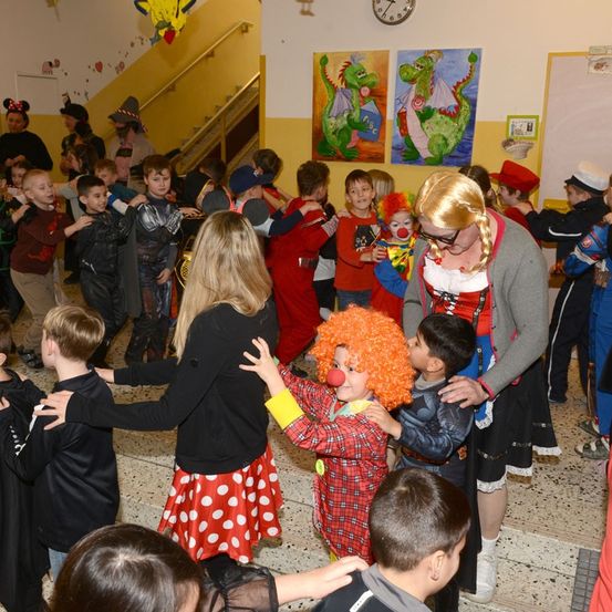Kinder in Kostümen tanzen in einem Raum mit Erwachsenen. Ein Clown unterhält die Kinder. Der Raum hat eine gelbe Wand und eine Treppe.