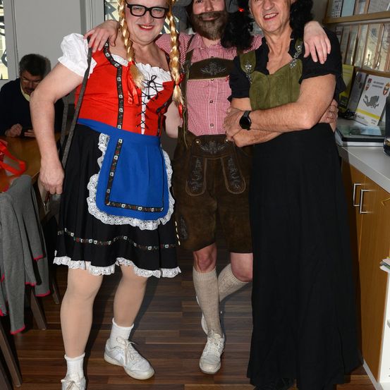 Drei Personen in traditioneller deutscher Kleidung stehen in einem Raum. Eine Frau in einem Dirndl, ein Mann in Lederhosen und eine ältere Frau in einem Kleid.