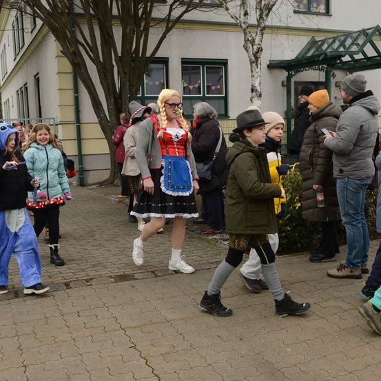 Eine Gruppe von Kindern und Erwachsenen geht vor einem Schulgebäude. Einige tragen Kostüme, andere lässige Kleidung. Die Kinder scheinen auf etwas zu warten.