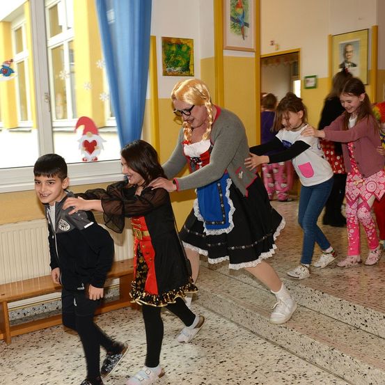Mehrere Kinder tanzen mit einer Frau in einem Kostüm. Der Raum hat gelbe Wände, und es gibt eine Bank in der Nähe eines Fensters.