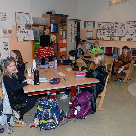Ein Klassenzimmer mit mehreren Kindern, die um Tische herum sitzen, ein Mädchen in einem roten Punktrock steht und ein Mädchen in einer Hexen-Kostüm liest aus einem Buch.