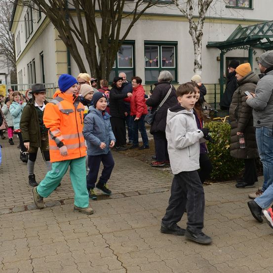 Eine Gruppe von Kindern und Erwachsenen geht auf einem Ziegelweg vor einem Gebäude. Einige tragen Winterkleidung und Mützen. Ein Mann in einer orangefarbenen Sicherheitsjacke führt die Gruppe an.
