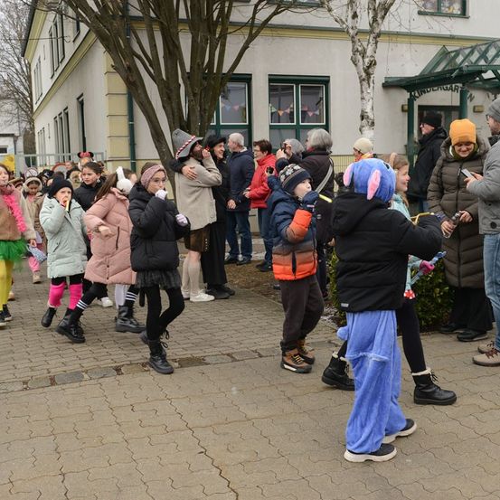 Eine Gruppe von Kindern und Erwachsenen in Winterkleidung, einige in Kostümen, versammelt sich vor einem Gebäude mit grünen Fenstern und einem Baum im Vordergrund.