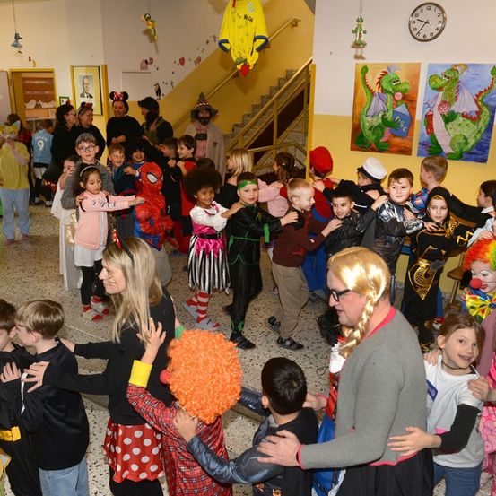 Eine Gruppe von Kindern in verschiedenen Kostümen, darunter Superhelden und Clowns, versammelt sich in einem Raum mit Erwachsenen. Sie scheinen an einer Kostümparty teilzunehmen. An der Wand hängen eine Uhr und zwei Gemälde.