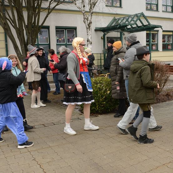 Eine Gruppe von Menschen in Kostümen vor einem Kindergarten. Einige benutzen Telefone, andere reden. Eine Frau in traditioneller Kleidung steht in der Mitte.