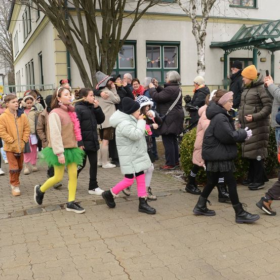 Eine Gruppe von Kindern und Erwachsenen in Kostümen geht die Straße entlang. Einige Kinder spielen Flöten und andere halten Kameras. Es gibt Bäume und ein Gebäude im Hintergrund.