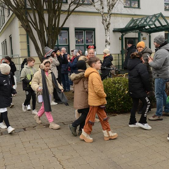 Eine Gruppe von Kindern und Erwachsenen in Kostümen und Jacken geht in einer Reihe vor einem Kindergartengebäude. Einige halten Kameras, während andere herumstehen und beobachten.