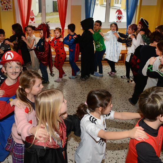 Eine Gruppe von Kindern in verschiedenen Kostümen steht in einem Kreis im Klassenzimmer, einige umarmen sich und andere tanzen. Sie scheinen an einem Halloween-Fest teilzunehmen.