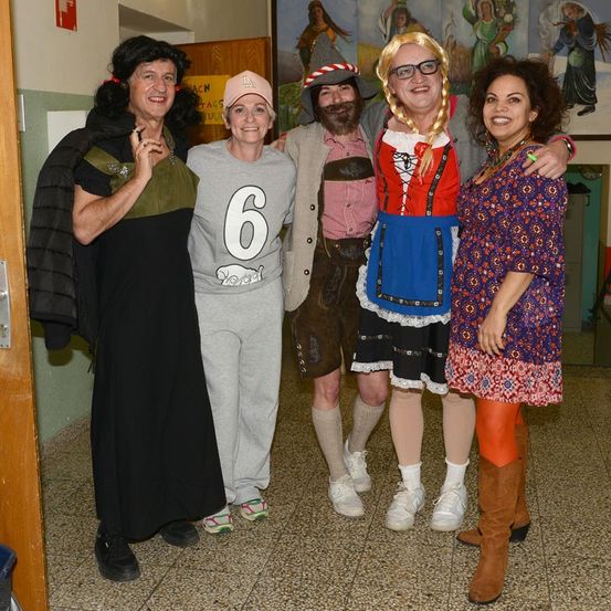 Fünf Personen in Kostümen stehen in einem Korridor. Eine Frau mit Bart und ein Mann mit Hut lächeln. Eine weitere Frau trägt ein '6' auf ihrem Shirt.