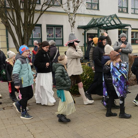 Eine Gruppe von Menschen in Winterkleidung, einige mit Mützen, gehen auf einem Ziegelweg. Im Hintergrund befindet sich ein Gebäude mit Glasfenstern.